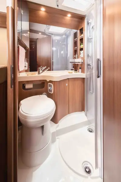 Das kompakte Wohnmobil-Badezimmer im Carthago C Tourer Teilintegriert verfügt über einen Holzschrank, eine weiße Toilette, ein kleines Waschbecken, einen Spiegel, ein Regal für Toilettenartikel, ein Handtuch und eine Eckdusche mit Glastür unter Deckenbeleuchtung.