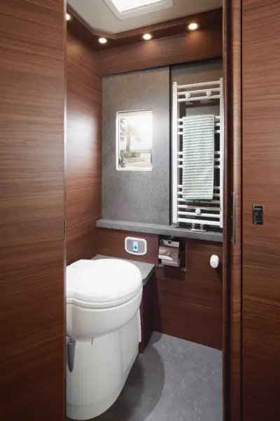 Ein kompaktes, modernes Badezimmer in der Carthago Chic C Line mit holzvertäfelten Wänden, einer weißen Toilette, einem kleinen Fenster, einem Handtuchhalter mit gefaltetem Handtuch, einem eingebauten Toilettenpapierhalter und einer Deckenbeleuchtung.