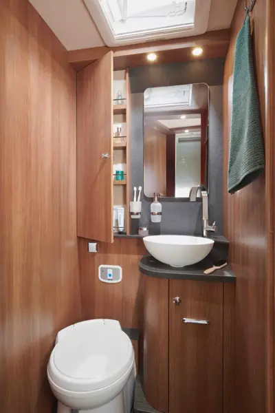 Ein kompaktes Badezimmer im Carthago C Tourer Teilintegriert, mit Holzvertäfelung, weißer Toilette, rundem Gefäßwaschbecken auf dunkler Arbeitsplatte, Spiegel, offenen Regalen mit Toilettenartikeln, Handtuchhalter und hellen Deckenleuchten sowie einem Oberlicht.