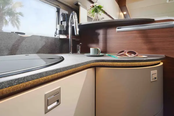 Moderne Wohnmobilküche in einem Carthago Chic C Line T mit geschwungener Arbeitsplatte, Spüle und eingebautem Herd. Eine Tasse, eine Sonnenbrille und ein Stift liegen auf dem Tresen, im Hintergrund blühen Blumen und das Sonnenlicht fällt durch das Fenster.