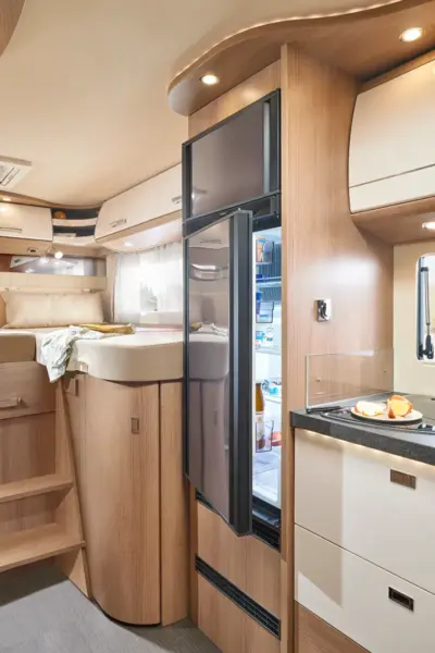 Modernes Wohnmobil-Interieur mit hellen Holzschränken, einem teilweise offenen Kühlschrank, einem kompakten Küchenbereich mit Theke und Herd, einer Treppe, die zu einem erhöhten Bett führt, und einem Fenster mit Vorhängen, die natürliches Licht hereinlassen - klassischer Carthago c tourer teilintegriert Stil.