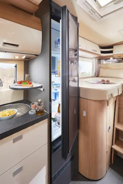 Die moderne Wohnmobilküche im Carthago C Tourer Teilintegriert verfügt über einen offenen Kühlschrank, frisches Gemüse auf einem ausziehbaren Tablett, Pommes frites, Gewürze auf der Theke, sowie ein gemütliches Bett und ein Fenster über einer Holztreppe im Hintergrund.