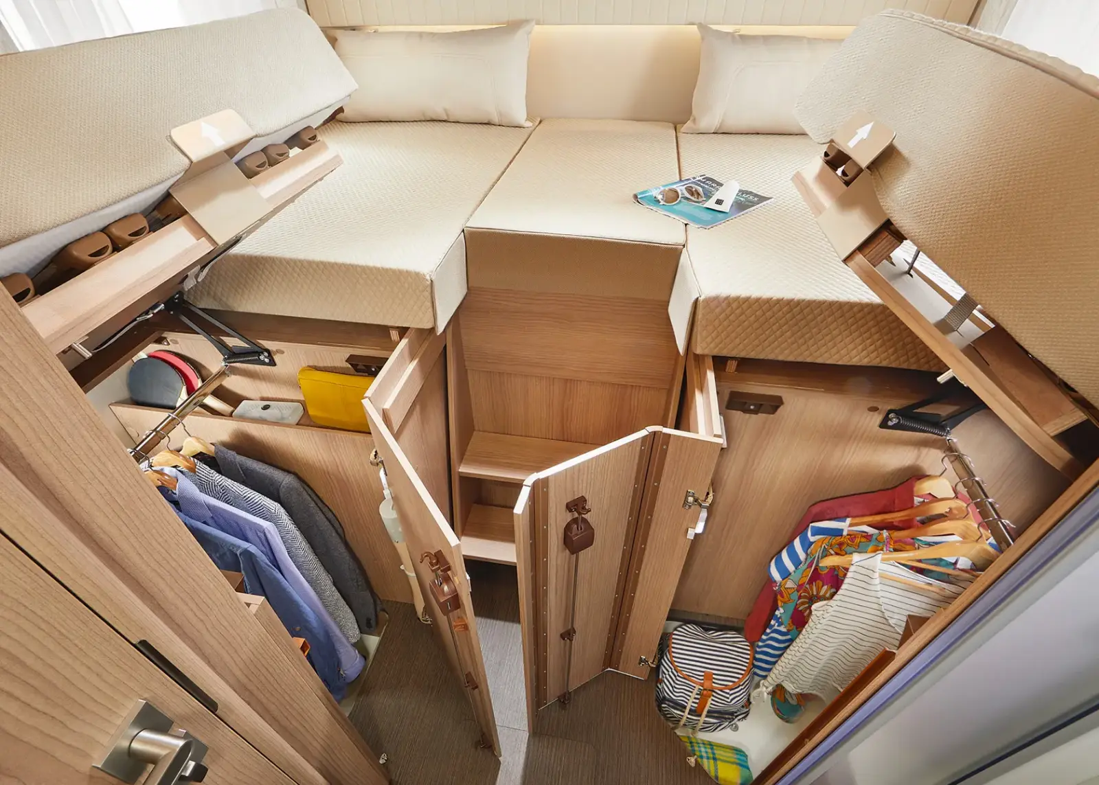 Offene Staufächer unter einem Bett in diesem Carthago C Tourer Teilintegriert offenbaren ordentlich organisierte Kleidung, Schuhe und Taschen. Auf dem beigen Bett liegen zwei Sonnenbrillen und ein Buch. Der kompakte Raum wird geschickt genutzt.