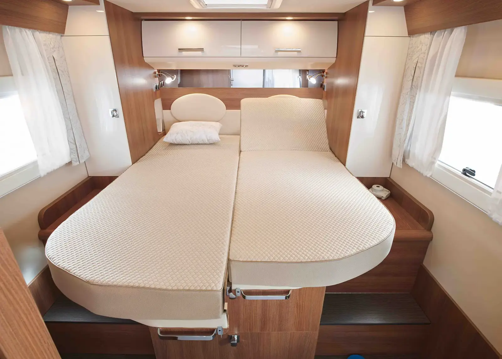 Zwei Einzelbetten mit beigen Matratzen und einem weißen Kissen in einem kompakten, modernen Carthago C Tourer Teilintegriert Wohnwagen-Schlafzimmer mit holzgetäfelten Wänden, eingebauten Schränken darüber und Seitenfenstern mit weißen Vorhängen, die Licht hereinlassen.