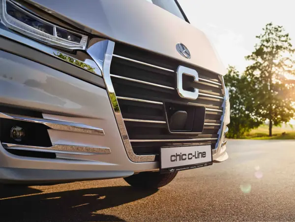 Nahaufnahme des Kühlergrills und der Stoßstange eines silbernen Carthago Chic C-Line, mit einem Chic C-Line-Kennzeichen, geparkt auf einer Straße mit Bäumen und Sonnenlicht im Hintergrund.