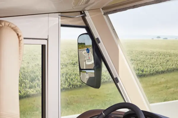 Blick aus dem Inneren eines Carthago Liner for Two, der durch ein Fenster auf einen Seitenspiegel blickt, in dem sich ein entferntes Kreisverkehrsschild spiegelt. Der Hintergrund zeigt ein grünes Feld unter einem leichten, dunstigen Himmel.
