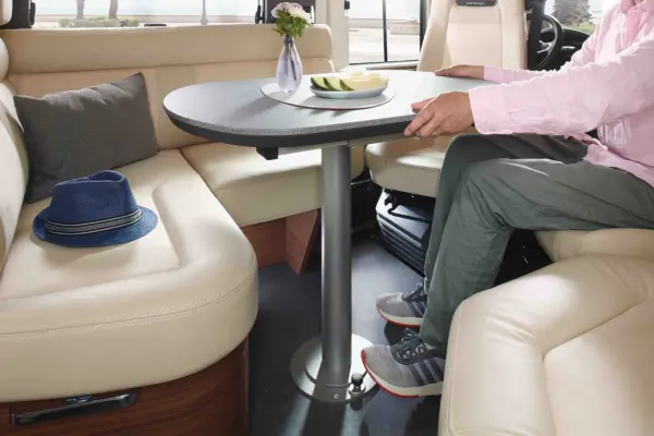 Eine Person in einem rosa Hemd sitzt an einem runden Tisch in einem modernen Carthago Chic C Line T Wohnmobil mit cremefarbenen Ledersitzen. Auf dem Tisch stehen eine kleine Vase mit Blumen, ein Tablett mit Essen und ein blauer Hut auf dem Sitz.