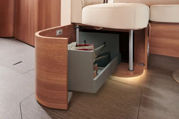 Ein Holzsofa mit eingebauter, ausziehbarer Schublade unter der Sitzfläche, ähnlich wie die clevere, platzsparende Konstruktion in einem Carthago C Tourer Teilintegriert. Eine sanfte Beleuchtung erhellt den Bereich um die mit Taschen gefüllte Schublade.