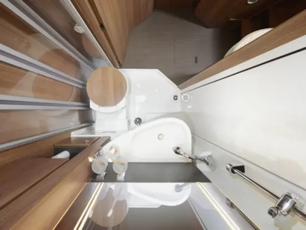 Draufsicht auf ein kompaktes Badezimmer in einem Carthago C2-Tourer mit einem runden Holz-WC-Sitz, einem kleinen weißen Waschbecken und einem Duschbereich, die alle nahtlos in einen schmalen Raum mit Holz und weißen Oberflächen eingepasst sind.