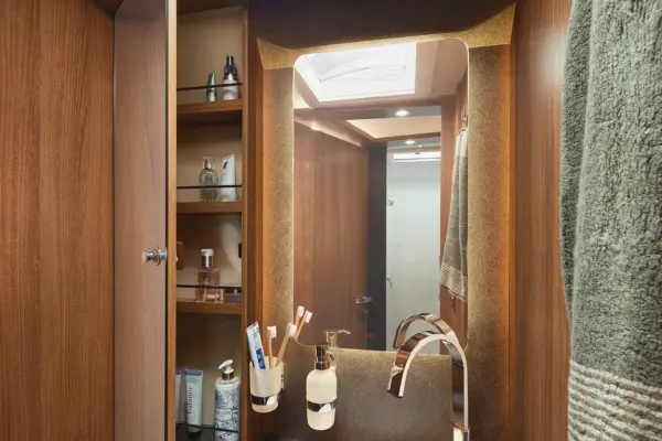 Das moderne Badezimmer im Carthago C2-Tourer verfügt über Holzschränke, offene Regale für Toilettenartikel, einen großen Spiegel, ein Handtuch auf der rechten Seite und einen Tresen mit einem Wasserhahn und Becherhaltern für Zahnbürsten und Zahnpasta.