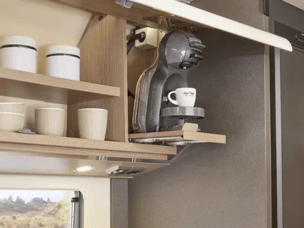 Eine carthago c2-tourer Kaffeemaschine mit einer Tasse steht auf einem ausziehbaren Regal in einem modernen Küchenschrank. In den benachbarten Regalen stehen weiße Tassen und Behälter, und ein Fenster darunter lässt natürliches Licht herein.