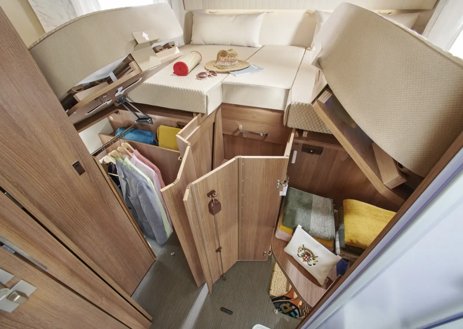 Ein gemütliches Carthago C2-Tourer Wohnmobil-Schlafzimmer mit einem erhöhten Bett, offenen Holzschränken, hängender Kleidung, gefalteten Handtüchern und persönlichen Gegenständen wie Hut, Buch und Sonnenbrille auf dem Bett.