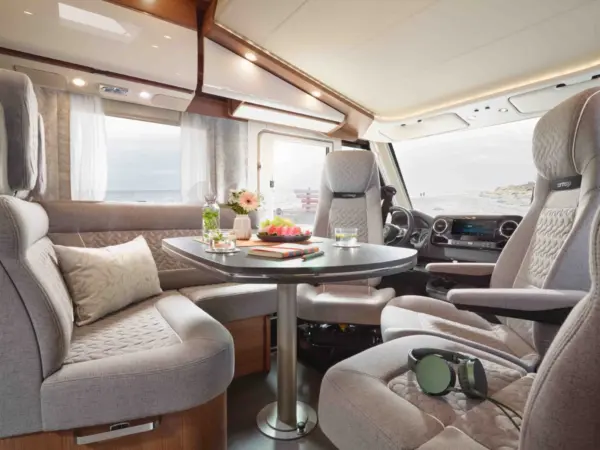 Ein modernes Carthago C2-Tourer Wohnmobil mit hellgrauen gepolsterten Sitzen, einem runden Tisch mit Snacks und Getränken, einem Fenster mit durchsichtigen Vorhängen, Blumen, Büchern und Kopfhörern auf dem Vordersitz.