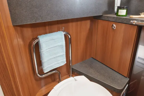 Ein modernes Badezimmer im Carthago Liner for Two verfügt über eine Holzverkleidung, eine weiße Toilette, einen verchromten Handtuchhalter mit einem hellblauen Handtuch und eine Arbeitsplatte mit einer grünen Kerze und einer Bürste.