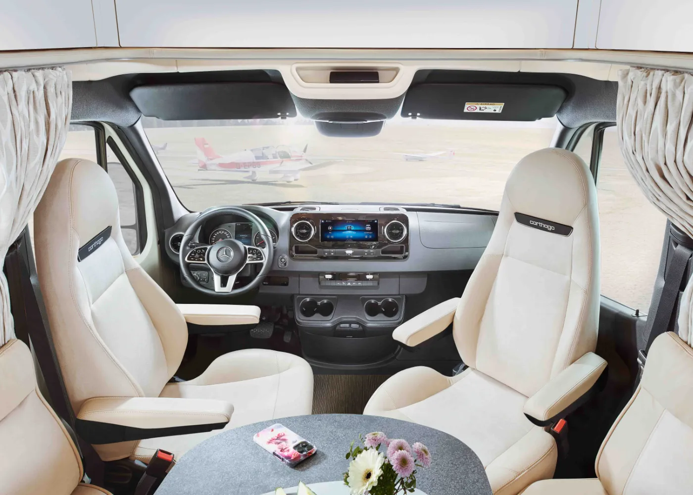 Innenansicht eines teilintegrierten Wohnmobil-Cockpits mit cremefarbenen Sitzen, zentralem Display, rundem Tisch mit Telefon und Blumen und zwei kleinen Flugzeugen, die durch die Windschutzscheibe sichtbar sind.