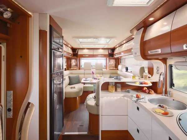 Modernes Interieur des Carthago Liner for Two mit schlichten Holz- und weißen Schränken, einer geschwungenen Küchentheke, Spüle, Herd und Kühlschrank. Es gibt einen gemütlichen U-förmigen Essbereich für eine Mahlzeit, mit Fenstern, die natürliches Licht hereinlassen.
