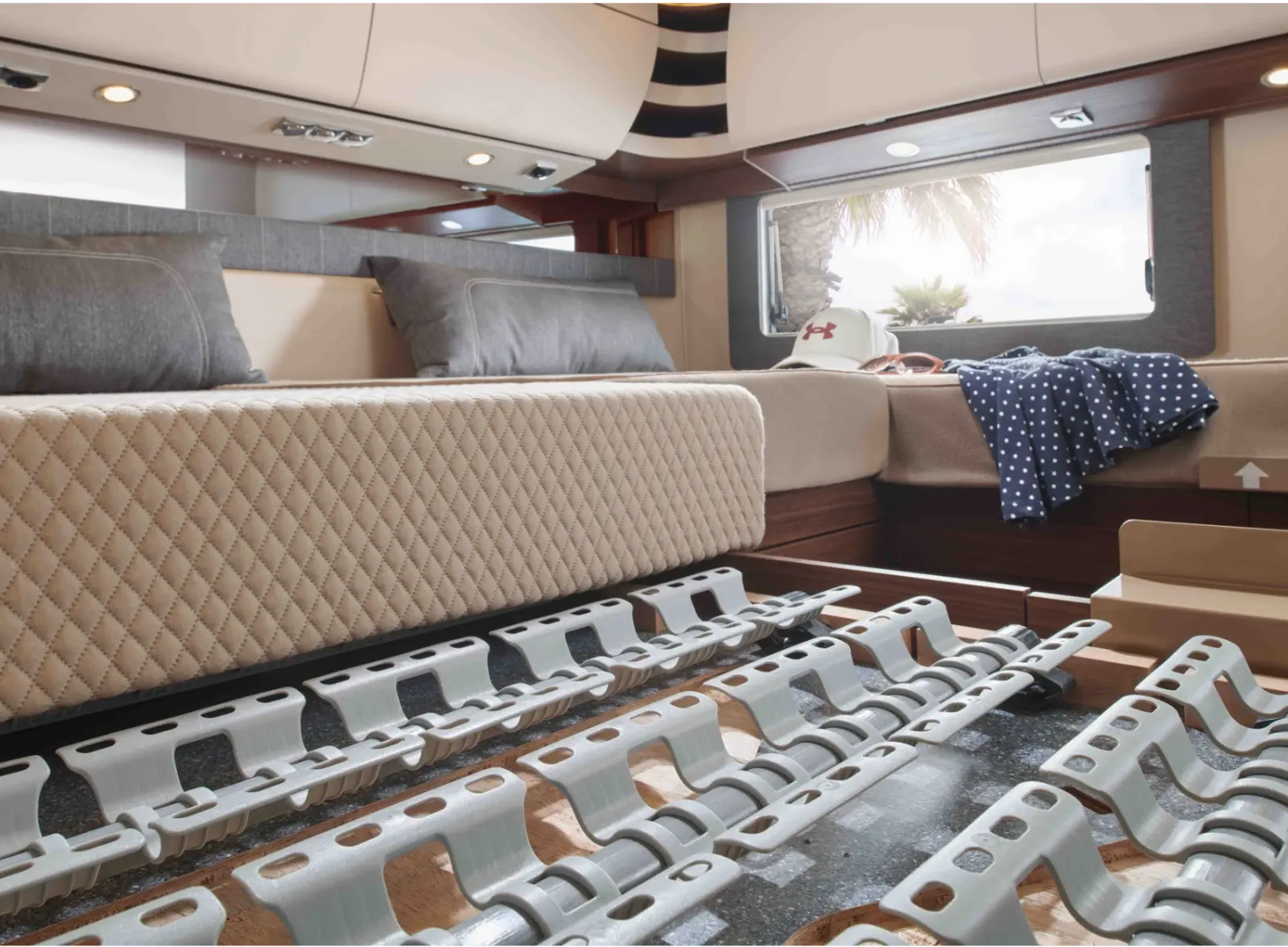 Eine Nahaufnahme eines modernen teilintegrierten Wohnmobils zeigt einen Bettrahmen mit einem angehobenen Lattenrost, der Stauraum darunter freigibt. Das Zimmer verfügt über neutrale Bettwäsche, Kissen, ein Fenster und einen Hut und ein gepunktetes Hemd auf dem Bett auf der anderen Seite.