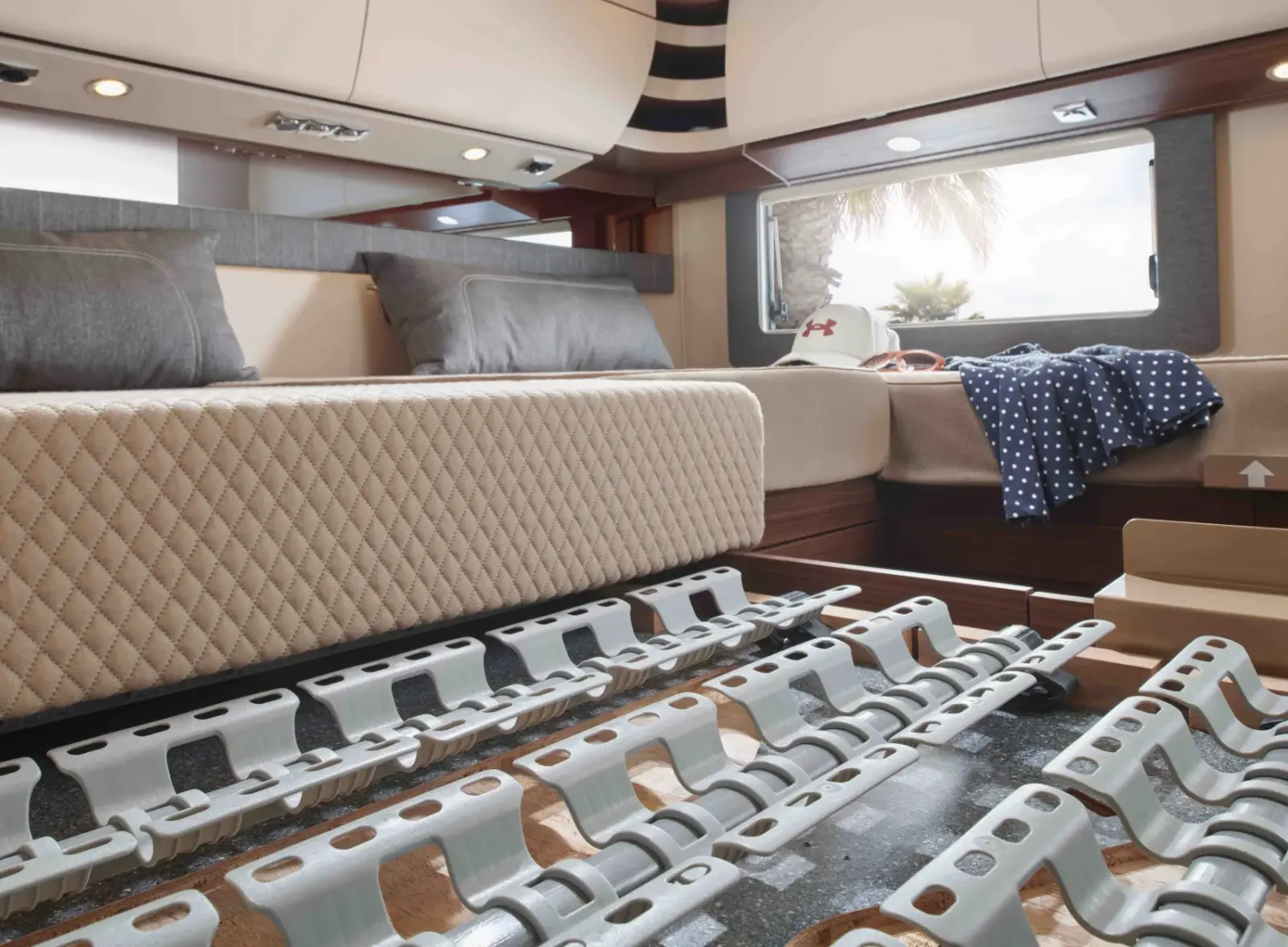 Innenansicht des Wohnmobil-Heckbereichs mit Fokus auf das ergonomische Schlafsystem. Die cremefarbene, rautengesteppte Matratze des großen Doppelbetts ist teilweise angehoben, um das darunterliegende Punktfederungssystem freizulegen. Viele einzelne, hellgraue Kunststoff-Federelemente sind auf Holzlatten befestigt und bilden eine flexible Unterkonstruktion. Im Hintergrund sind Kissen, Oberschränke und ein Fenster mit Blick auf Palmen zu sehen, was die komfortable Reiseatmosphäre unterstreicht.