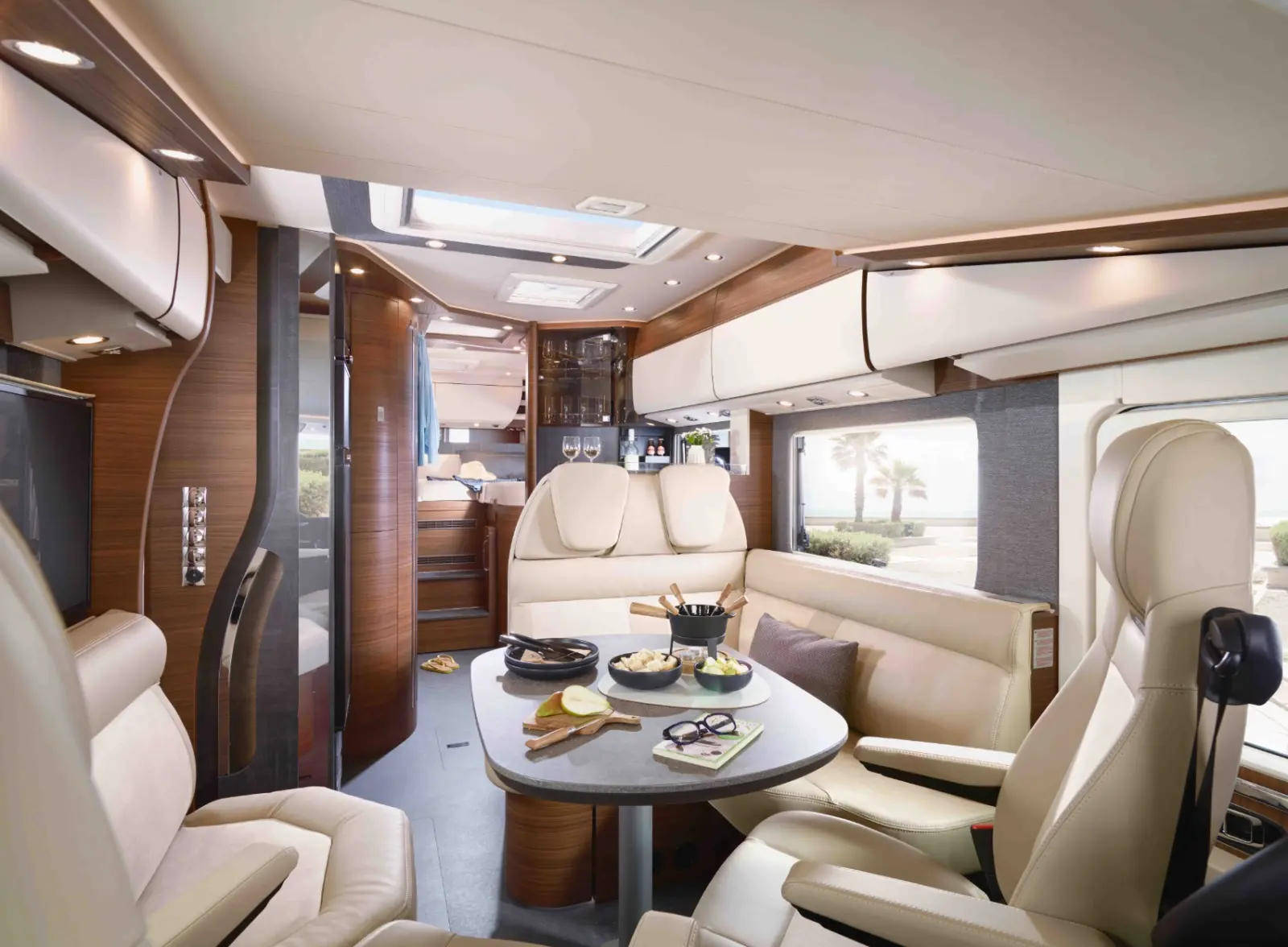 Innenraum eines luxuriösen vollintegrierten Wohnmobils mit cremefarbenen Ledersitzen, einem runden Tisch mit Snacks und einem Blick auf Palmen und das Meer durch große Fenster. Der Raum wirkt hell, modern und komfortabel.