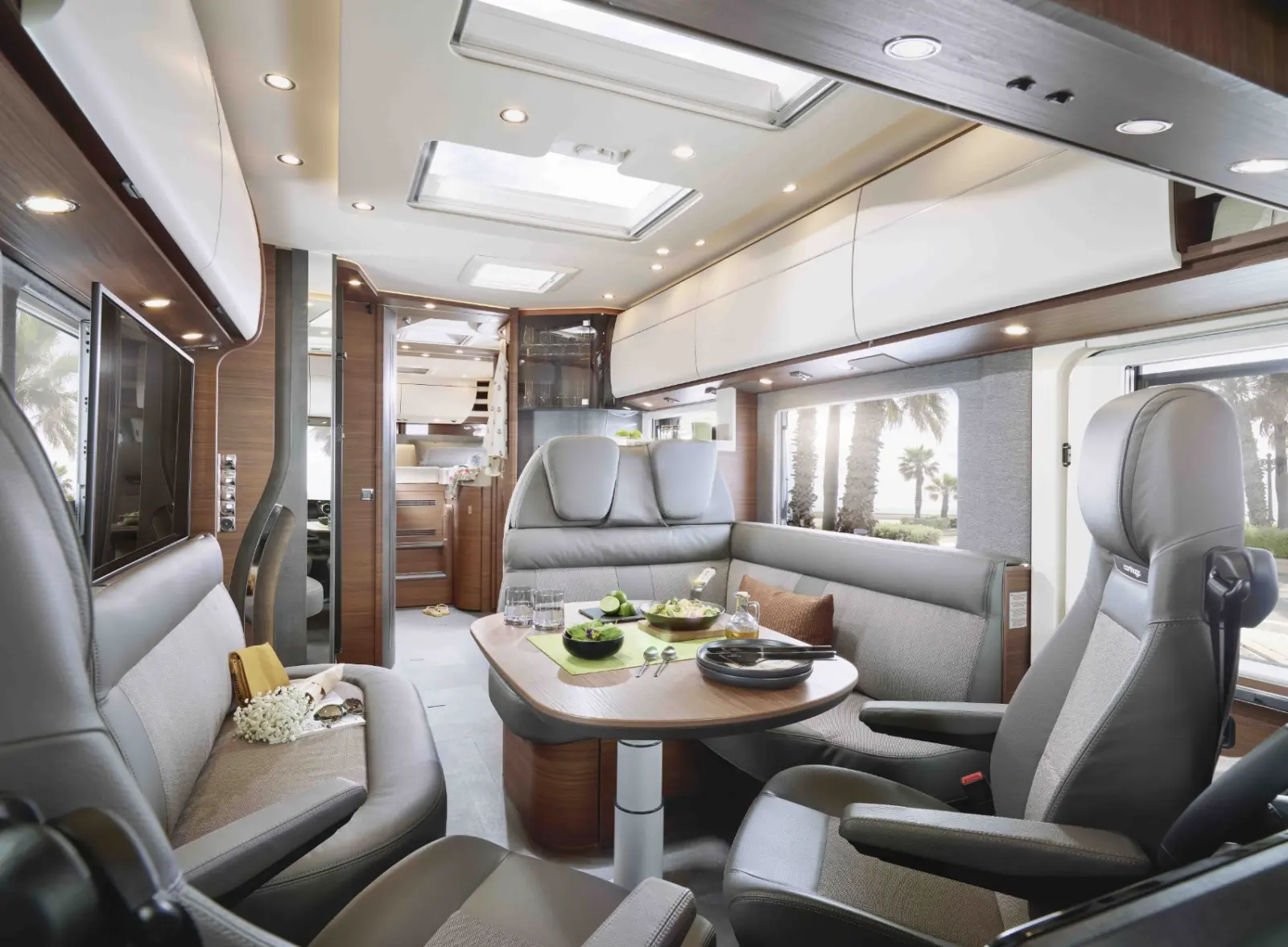 Modernes vollintegriertes Wohnmobil-Interieur mit glatten grauen Sitzen und hölzernen Akzenten, einem Esstisch mit Tellern und Getränken, Stauraum über der Decke, Deckenfenstern und einem Blick auf Palmen durch das Seitenfenster.