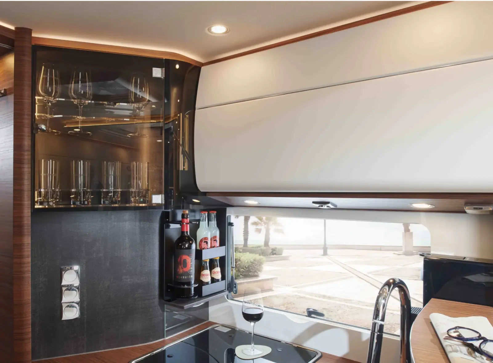 Eine moderne Wohnmobilküche verfügt über eine Glasvitrine mit Weingläsern, ein ausziehbares Regal mit Weinflaschen und Dosengetränken, ein Glas Rotwein auf dem Tresen und ein Fenster mit Blick aufs Meer.