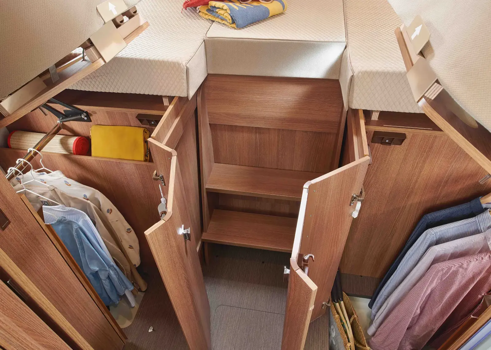 Offener Holzschrank mit Regalen und hängender Kleidung, einschließlich Hemden und Jacken, in einem ordentlich organisierten Raum in einem Carthago C Tourer Teilintegriert. Darüber befindet sich ein Bett mit gefalteter Kleidung, was auf einen gemütlichen, kompakten Wohnbereich hindeutet.