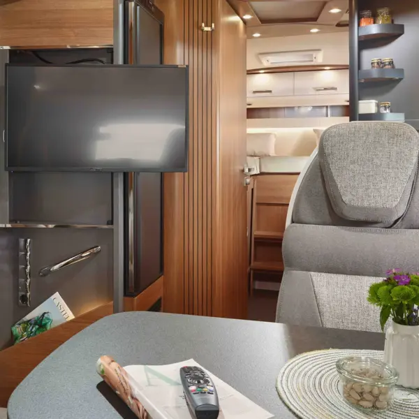 Innenraum eines modernen Carthago C Tourer Teilintegrierten Wohnmobils mit eingebautem Fernseher, grauem Polstersitz, Tisch mit einer Zeitschrift, Fernbedienung, Blumenvase und Glasgefäß. Im Hintergrund sind ein gemütliches Bett und Regale mit Gläsern zu sehen.