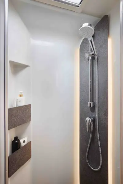 Moderne Dusche mit großer Regendusche, Handbrause, zwei eingelassenen Ablagen für Flaschen und einem dunklen vertikalen Akzentpaneel vor weißen Wänden - kompakt und lichtdurchflutet, in Anlehnung an das raffinierte Design der Carthago Chic C Line T.