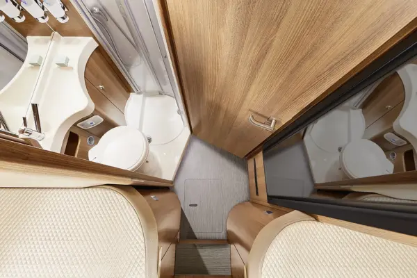 Draufsicht auf ein kompaktes Wohnmobil-Badezimmer des Carthago C Tourer Teilintegriert mit Holzvertäfelung, weißer Toilette, kleinem Winkelwaschbecken und Spiegelschrank, angrenzend an einen Sitzbereich mit strukturierter Polsterung.