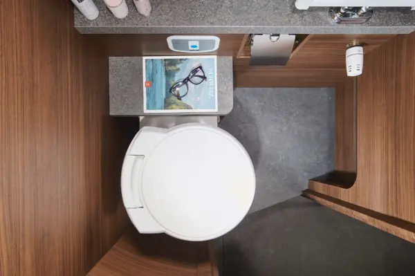 Draufsicht auf ein modernes Badezimmer in einem Carthago Chic C Line, mit einer geschlossenen weißen Toilette, einem Buch mit Lesebrille, holzgetäfelten Wänden, einem kleinen Regal mit Gegenständen und einer schlichten grauen Arbeitsplatte.