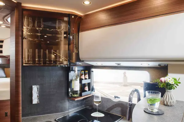 Eine moderne Wohnmobilküche im Carthago Chic C Line T ist mit einem Glasschrank, Weinflaschen, einem Waschbecken, einem Glas Weißwein, Wasser mit Limette und einer kleinen Blumenvase auf der Arbeitsplatte in der Nähe eines Fensters ausgestattet.