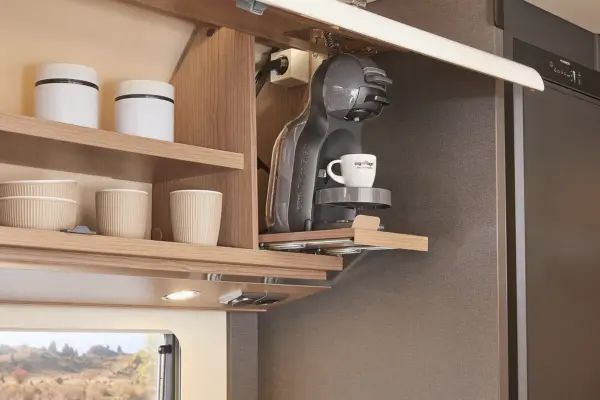 Ein moderner Küchenschrank in einem Carthago C Tourer Teilintegriert verfügt über ein offenes Regal mit Tassen und Behältern sowie eine eingebaute Kaffeemaschine mit einer Tasse auf dem Tablett, alles vor dem Hintergrund einer schlichten, farbneutralen Einrichtung.