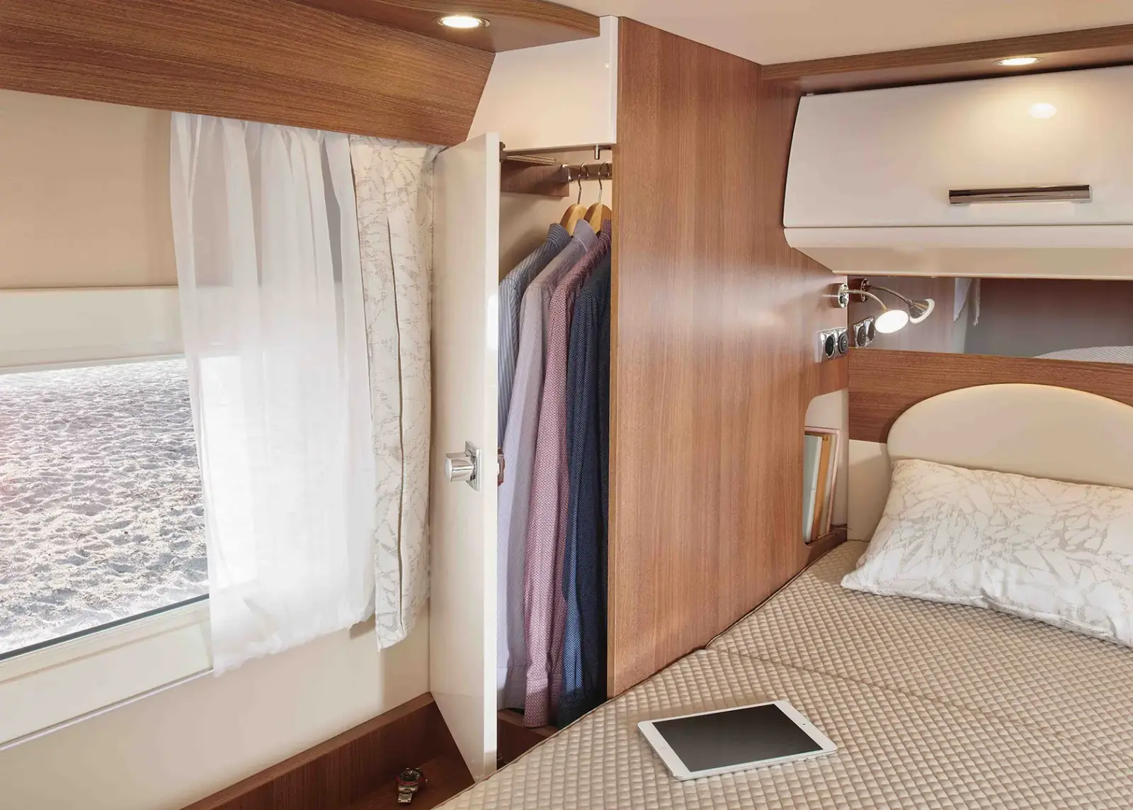 Ein gemütliches Carthago C Tourer Teilintegriert Wohnmobil mit einem gemachten Bett, einem Tablet auf der Decke, einem Fenster mit durchsichtigen Vorhängen und einem offenen Kleiderschrank mit ordentlich aufgehängter Kleidung auf Holzbügeln.