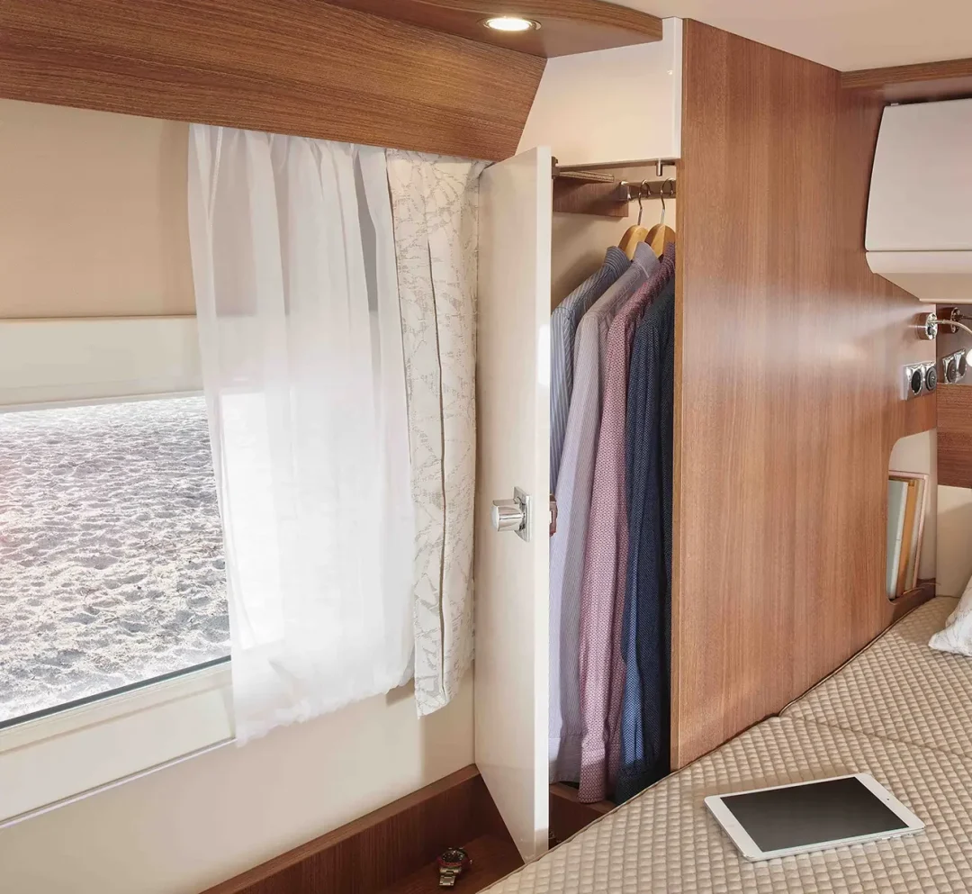 Ein gemütliches Carthago C Tourer Teilintegriert Wohnmobil mit einem gemachten Bett, einem Tablet auf der Decke, einem Fenster mit durchsichtigen Vorhängen und einem offenen Kleiderschrank mit ordentlich aufgehängter Kleidung auf Holzbügeln.