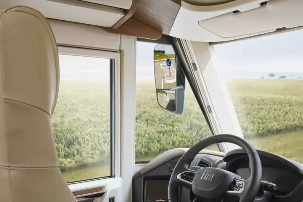 Blick aus dem Inneren eines Carthago Chic C-Line Wohnmobils mit Fahrersitz, Lenkrad, Fenster und Seitenspiegel, in denen sich ein Straßenschild spiegelt; draußen erstreckt sich ein grünes Feld unter einem bewölkten Himmel.