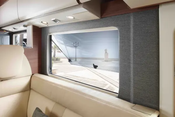 Blick aus dem Inneren eines Carthago Chic C Line T Wohnmobils, mit einem beigen Ledersofa, einer grauen Wand und einem offenen Fenster mit einem Bildschirm. Draußen sind eine sonnige Straße, Palmen und eine Statue zu sehen.