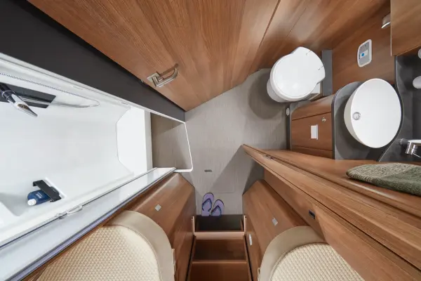 Draufsicht auf ein kompaktes, modernes Wohnmobil-Badezimmer in einem Carthago C2-Tourer mit Holzschränken, einem runden Waschbecken, einer kleinen Toilette, einer Dusche und einem beigen Bodenbelag. Ein Paar Hausschuhe und ein gefaltetes Handtuch sind zu sehen.