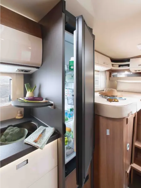 In der modernen Wohnmobilküche des Carthago C2-Tourer steht ein schmaler Kühlschrank offen und gibt den Blick frei auf ordentlich sortierte Lebensmittel. In der Nähe befindet sich eine Spüle mit frischem Gemüse, und in dem gemütlichen Holzinterieur ist ein Bett mit einem Hut darauf zu sehen.