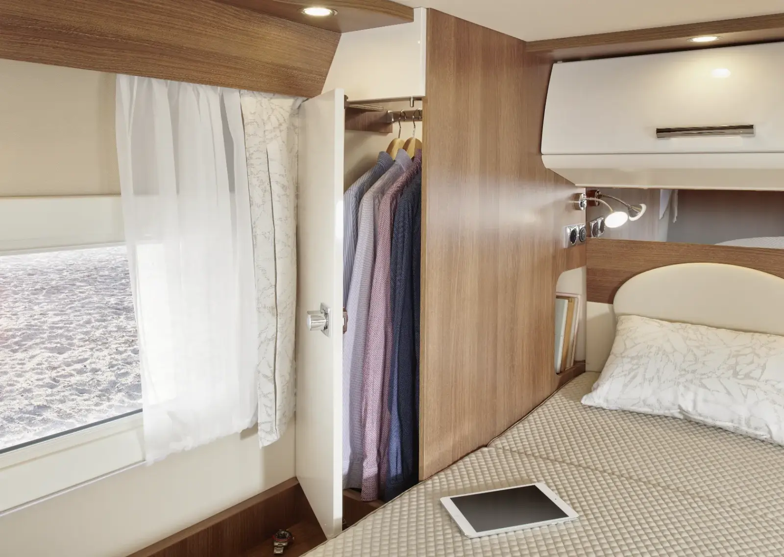 Ein aufgeräumtes Carthago C2-Tourer Wohnmobil-Interieur mit einem sauber gemachten Bett, einem offenen Kleiderschrank mit Hemden auf Bügeln, einem Fenster mit weißen Vorhängen und einem Tablet auf der Bettdecke.