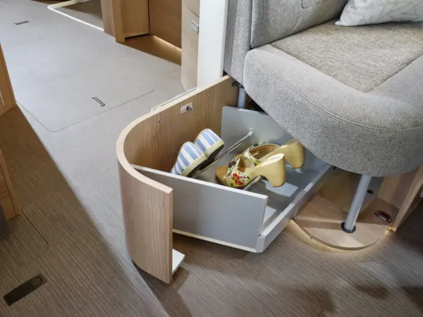 Eine in den Sockel eines beigen Sofas eingebaute Schublade bietet Platz für Damensandalen und Keilabsätze und sorgt so für Ordnung in einem modernen Carthago C2-Tourer Wohnmobil mit hellem Holzfußboden und hellem Mobiliar.
