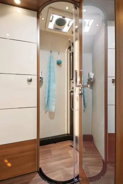 Eine moderne Dusche im Carthago Liner for Two mit einer gewölbten Glastür, Wänden aus Holz und weißen Paneelen, einer Regendusche an der Decke, einem blauen Handtuch im Inneren, Toilettenartikeln auf einem kleinen Regal und einem Fußboden mit Holzdekor.