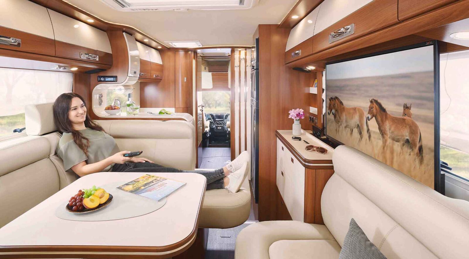 Eine Frau entspannt sich auf einem beigen Sofa in einem integrierten Wohnmobil und sieht sich eine Fernsehsendung über Wildpferde an. Auf dem Tisch stehen eine Schale mit Obst und eine Zeitschrift, umgeben von Holzschränken und stimmungsvoller Beleuchtung.