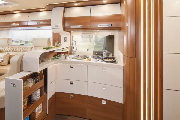 Moderne Wohnmobilküche in einem Carthago Liner for Two, mit glänzend weißen und hölzernen Schränken, eingebautem Herd, Spüle, Unterschrankbeleuchtung, offenen Regalen mit Lebensmitteln und einer gemütlichen Sitzecke im Hintergrund.