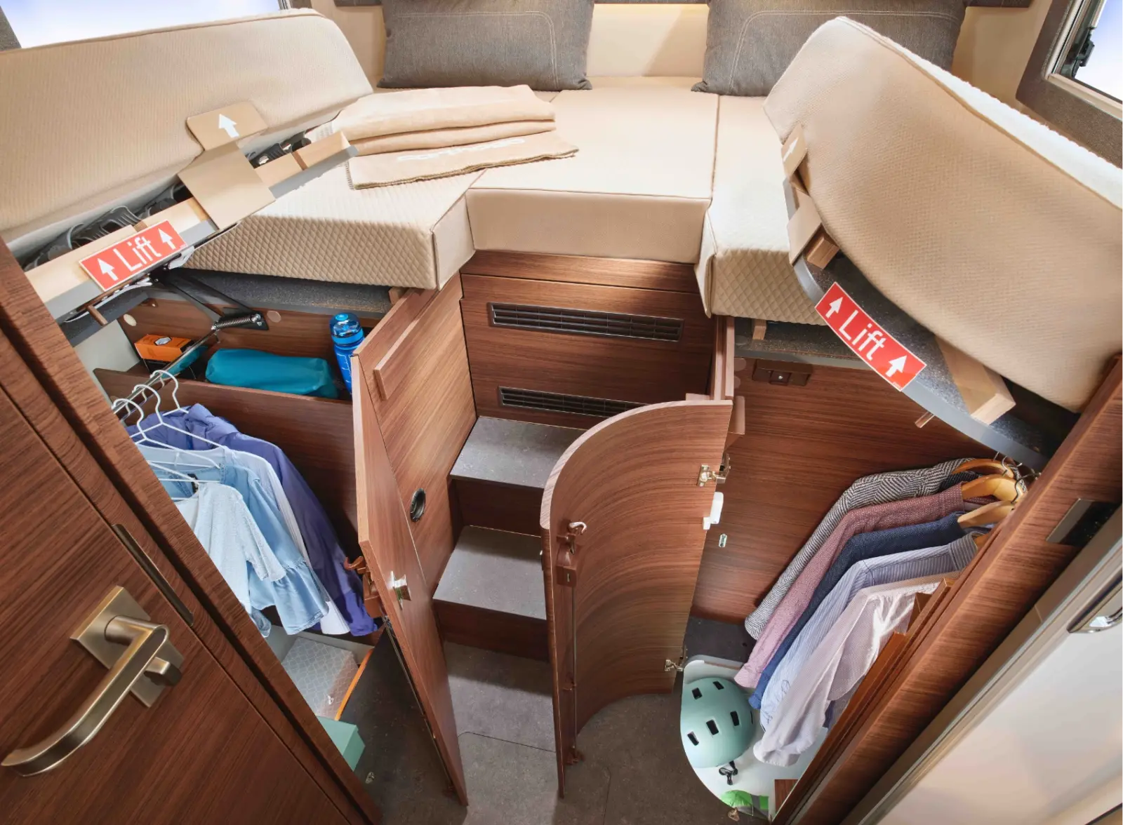 Innenraum eines integrierten Wohnmobils mit einem erhöhten Bett, das teilweise angehoben wird, um einen Schrank und Stauraum darunter freizugeben. Kleidung hängt an Regalen, eine Treppe führt nach oben, und rote Lift-Schilder weisen auf die Hebefunktion des Bettes hin.