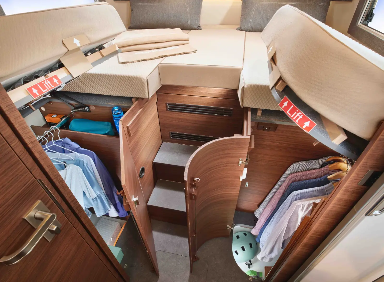 Offener Schrankraum unter einem Hochbett in einem integrierten Wohnmobil, mit hängenden Kleidungsstücken, einer Wasserflasche, Ablagefächern und einer Treppe, die nach oben führt, mit Pfeilen, die darauf hinweisen, dass das Bett angehoben werden kann.