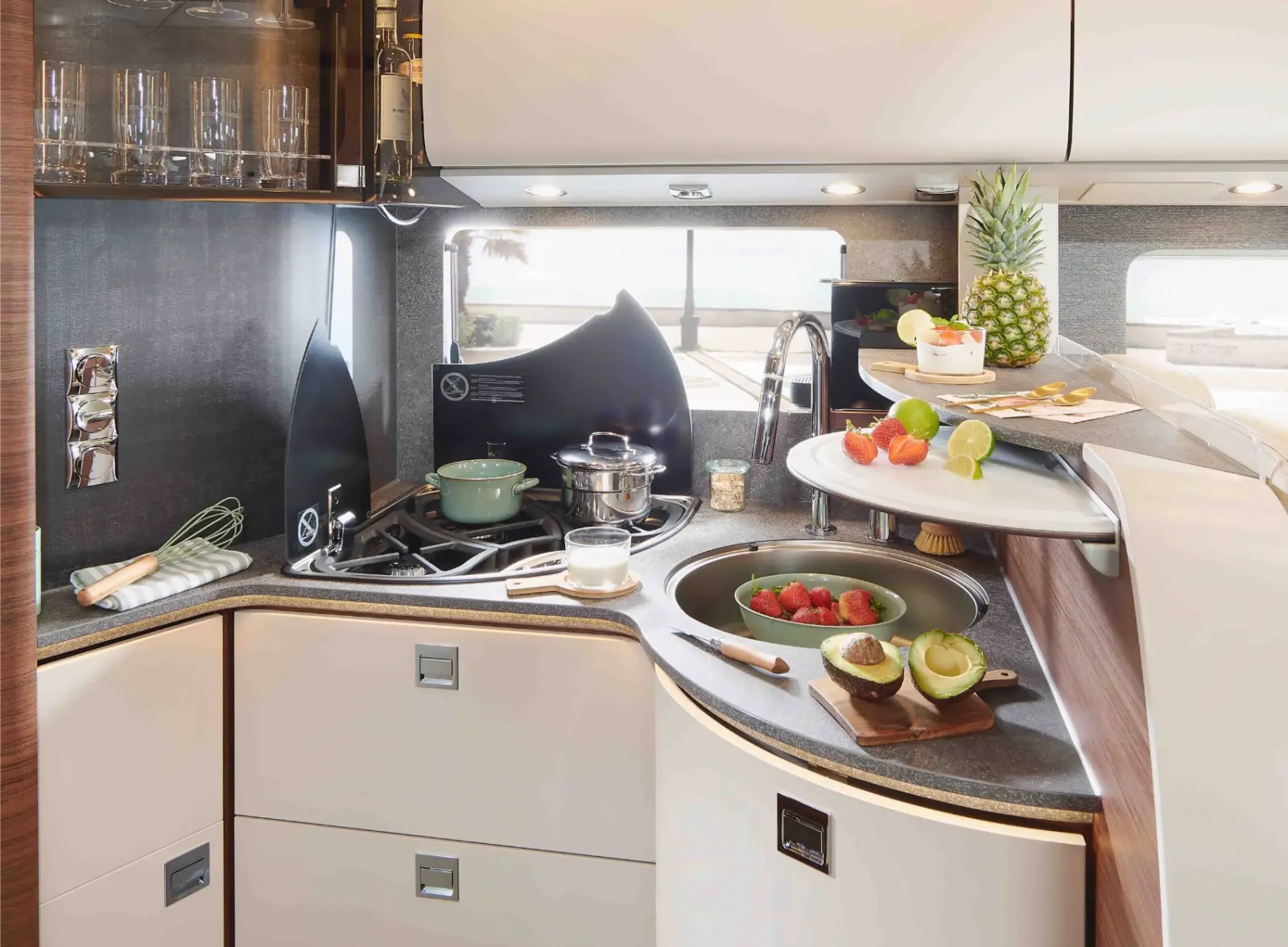 Moderne Wohnmobilküche mit weißen Schränken, einem Herd mit Töpfen, einer Spüle und frischen Früchten wie Erdbeeren und Avocado auf der Theke. Große Fenster füllen dieses vollintegrierte Wohnmobil mit reichlich natürlichem Licht für einen hellen, einladenden Raum.