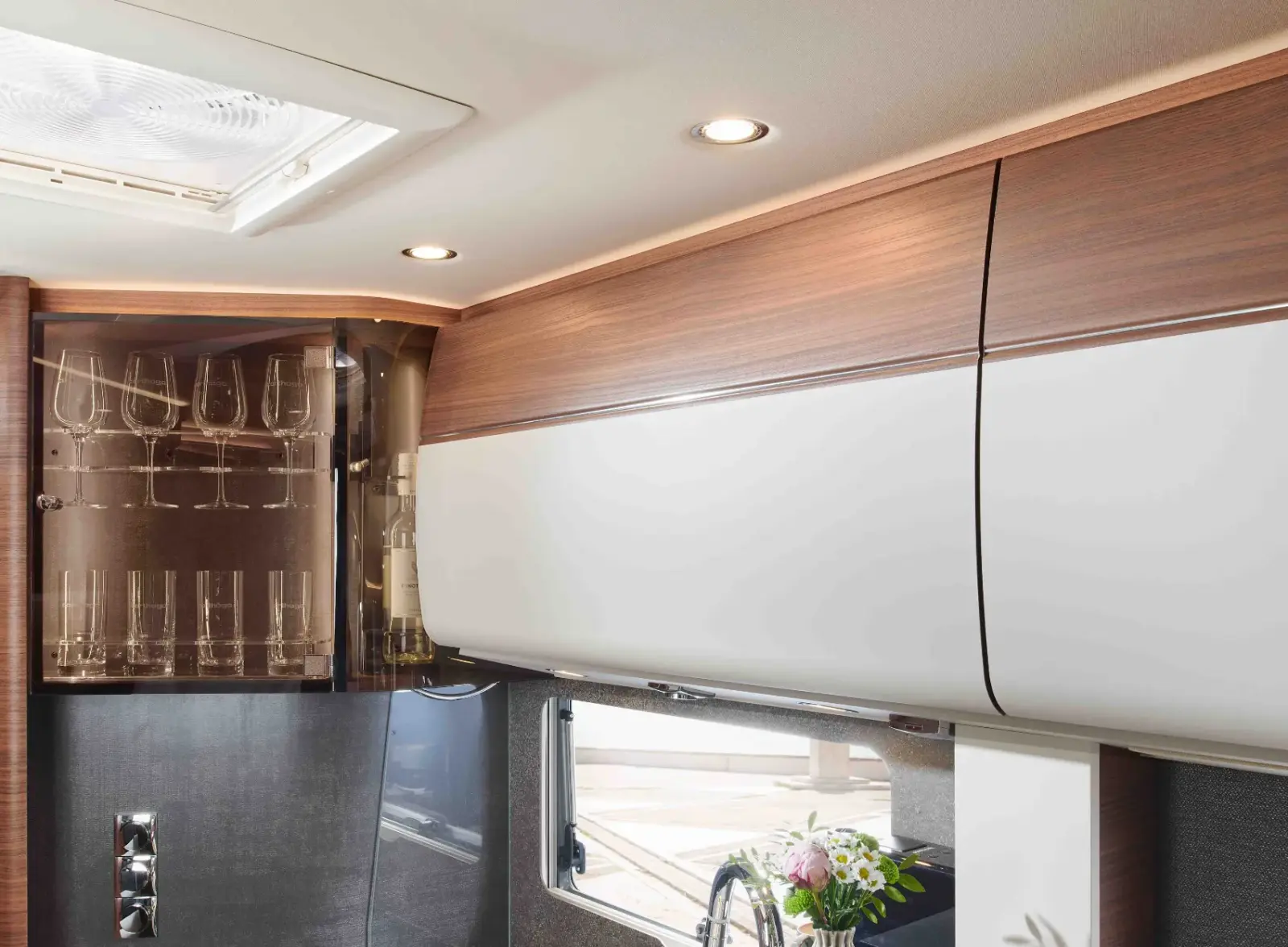 Modernes Wohnmobil-Interieur mit Schränken in Holz und Weiß, eingebautem Glaswarenregal mit umgedrehten Weingläsern, einem Fenster, in dem sich Blumen spiegeln, und Deckenleuchten, die für eine warme Beleuchtung sorgen.