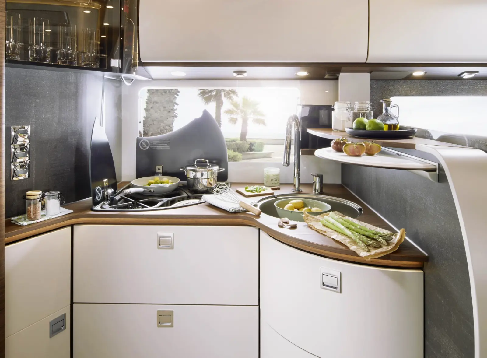 Moderne integrierte Wohnmobil-Küche mit glatten weißen Schränken, Herd, auf dem Essen gekocht wird, Spargel auf einem Schneidebrett, frisches Obst und Gemüse, Spüle mit fließendem Wasser und ein Fenster mit Palmen und Sonnenlicht draußen.