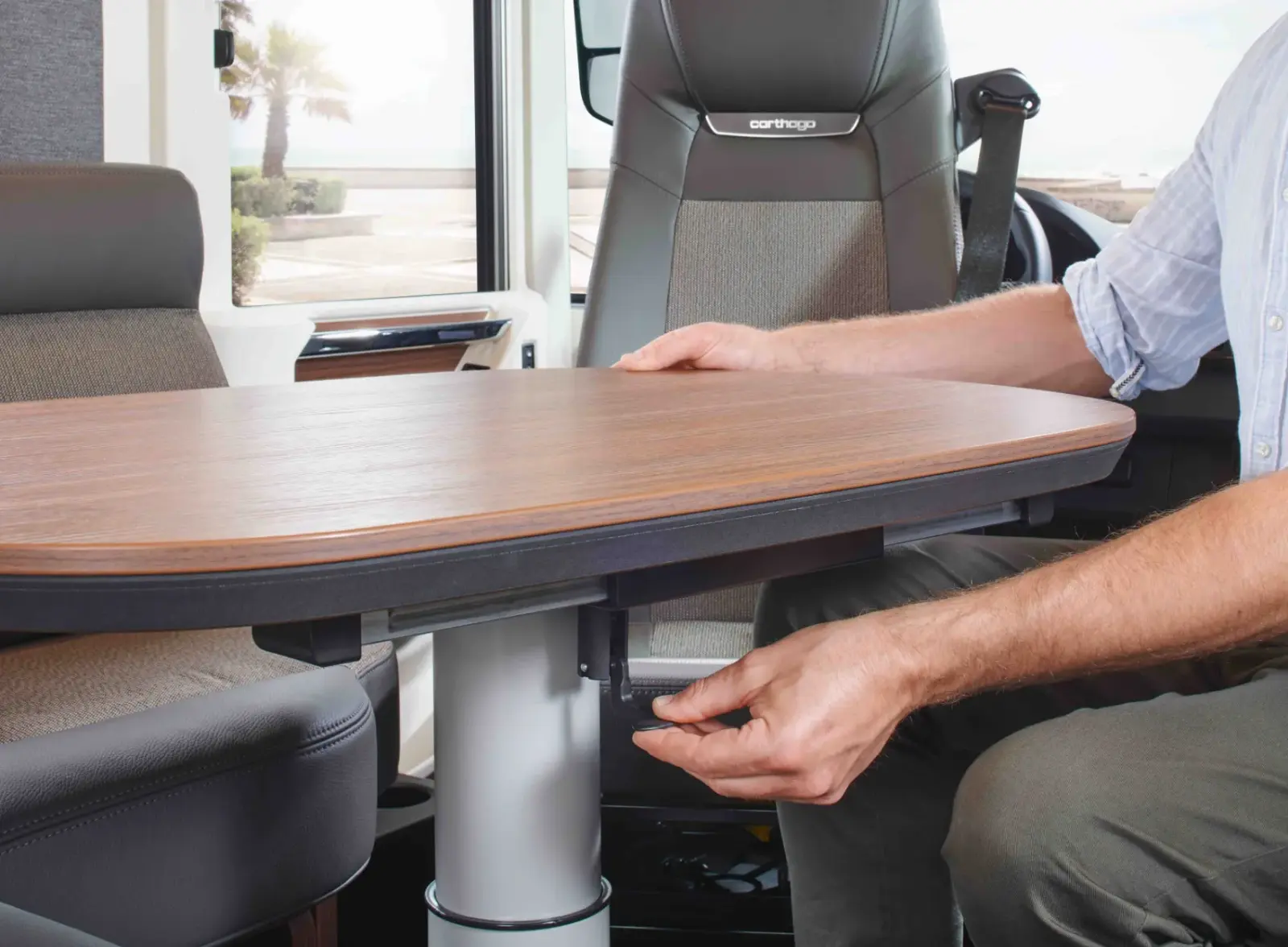 Eine Person verstellt die Höhe eines Holztisches in einem modernen Fahrzeug oder Wohnmobil. Der Tisch ist an einem Metallgestell befestigt, und im Hintergrund sind Sitze zu sehen.