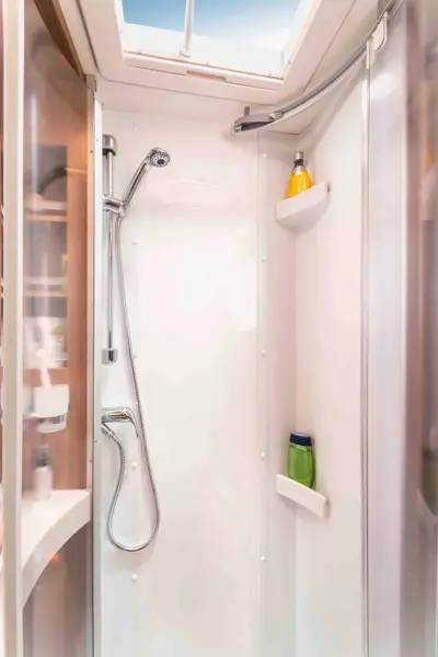 Die kompakte, moderne Dusche im Carthago C Tourer Teilintegriert verfügt über eine Handbrause, weiße Wände, zwei Eckablagen für Flaschen und ein Oberlicht, das natürliches Licht hereinlässt.